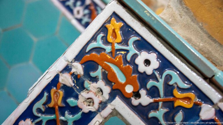 Close-up of chipped blue glazed tile with orange palmettes, white flowers, and turquoise scrolls.