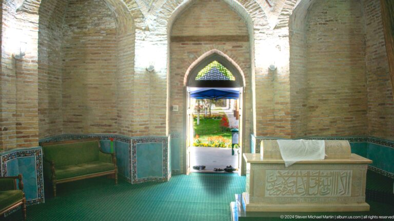 Green-carpeted mausoleum interior with pointed doorway to a bright garden and cenotaph at right.