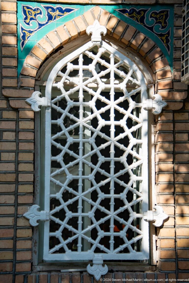 White geometric metal grille covering a narrow opening in a tan brick wall.