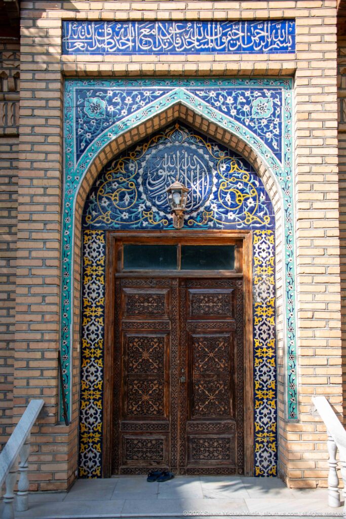 Carved wooden door beneath blue floral tilework and white Arabic calligraphy in a pointed arch.