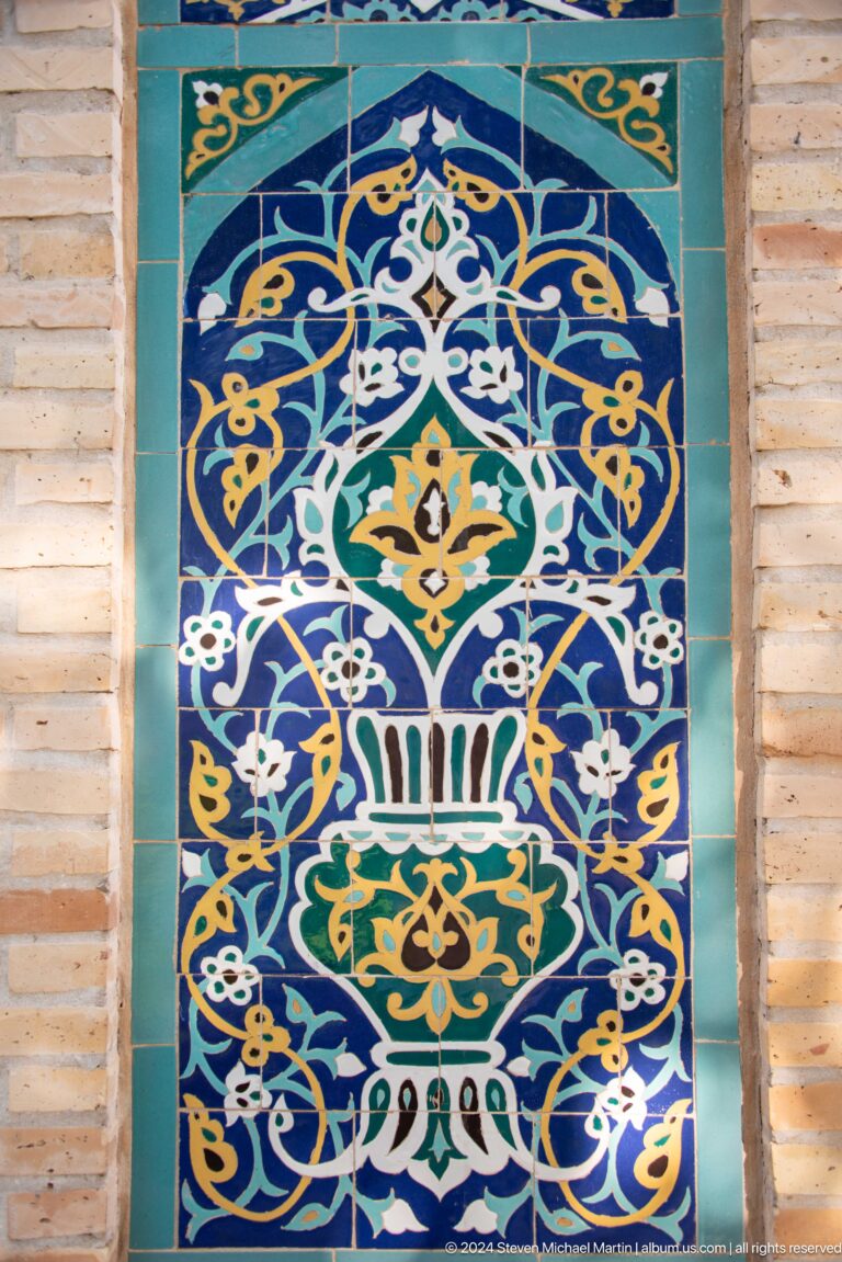 Blue glazed tile panel with a white-and-gold vessel, flowers, and symmetrical curling vines.