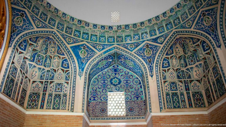 Curved mausoleum wall covered with blue tiled pointed niches around a glowing lattice window.