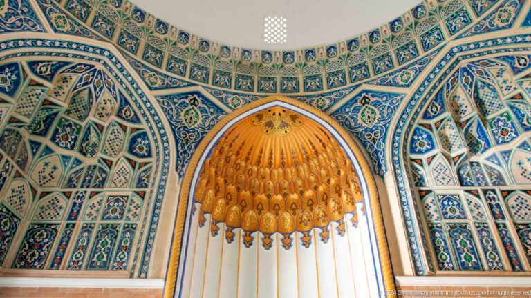Golden muqarnas canopy above a tall white curtain, framed by blue patterned tiles.