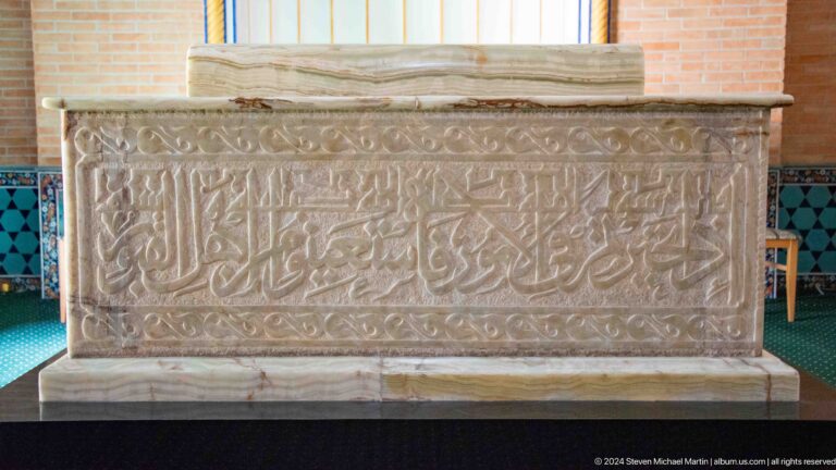 Arabic inscription carved across the side of a pale stone cenotaph beneath a white cloth.