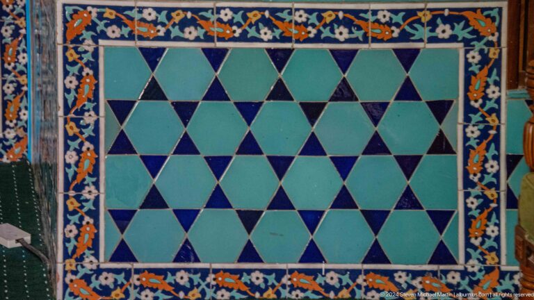 Rectangular tile panel of turquoise and dark blue triangles framed by a floral border.