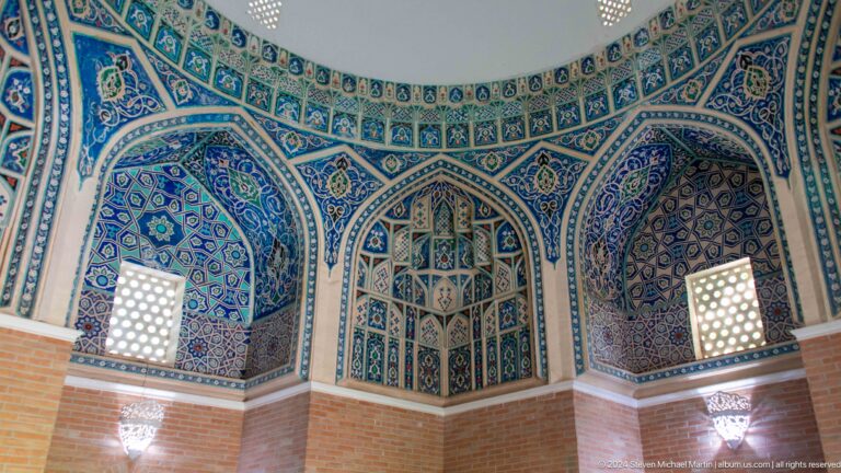 Wide mausoleum interior with three blue tiled niches, two glowing lattice windows, and brick lower walls.