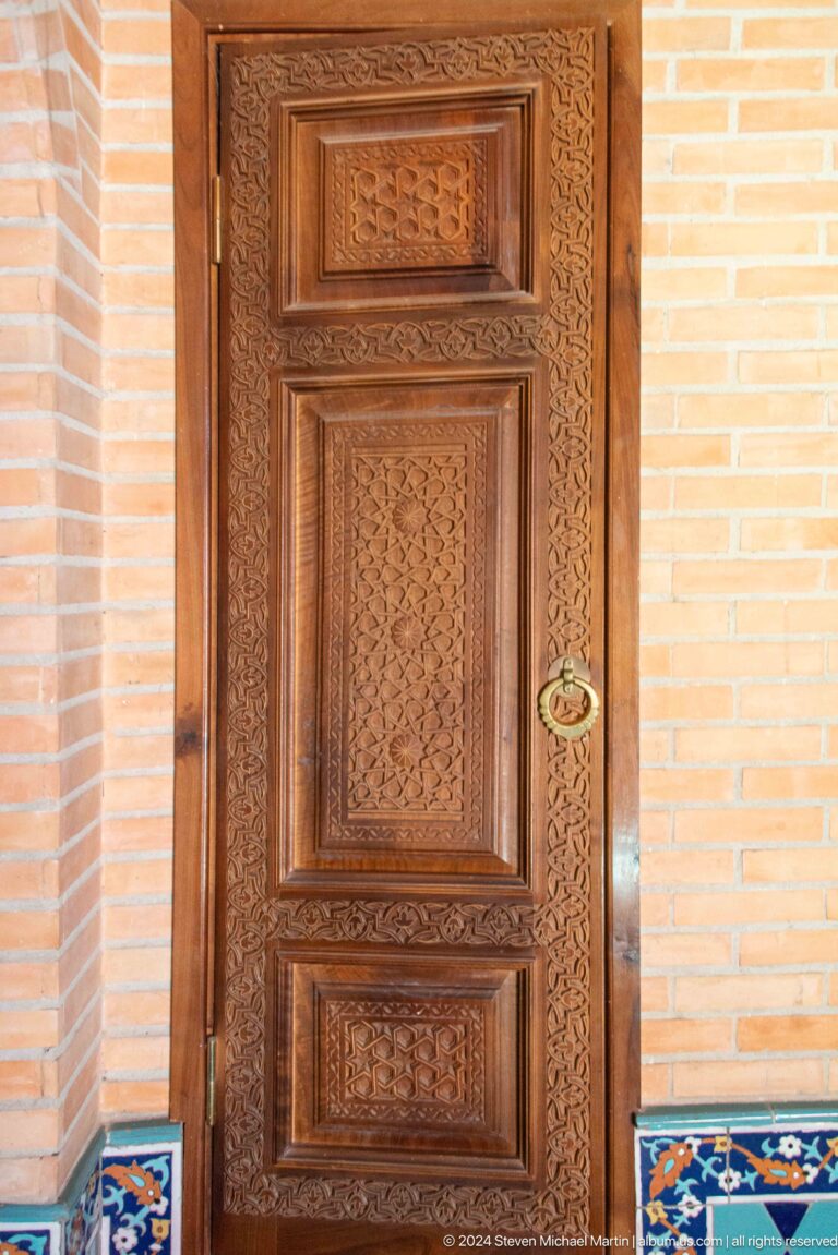Carved single wooden door with dense ornament and brass ring pull in a tan brick recess.
