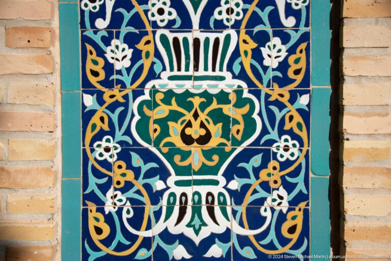 Blue glazed tile panel with a white urn, green medallion, gold vines, and turquoise leaves.