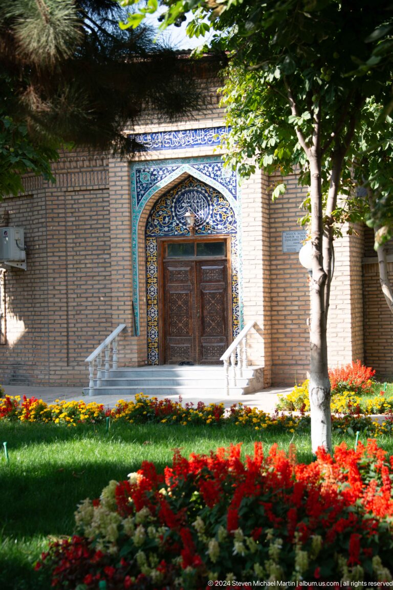 Blue-tiled pointed doorway with carved wood door, white steps, flowers, and trees.