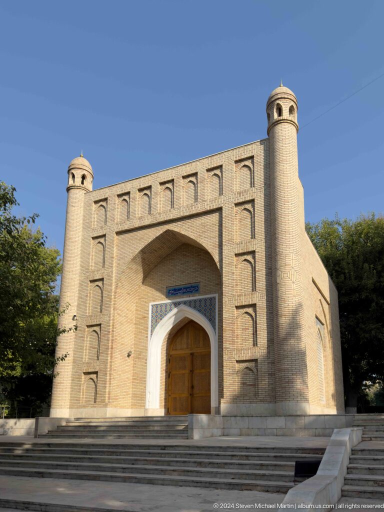 Frontal brick mausoleum facade with deep pointed portal, carved door, blue inscription, and corner turrets.