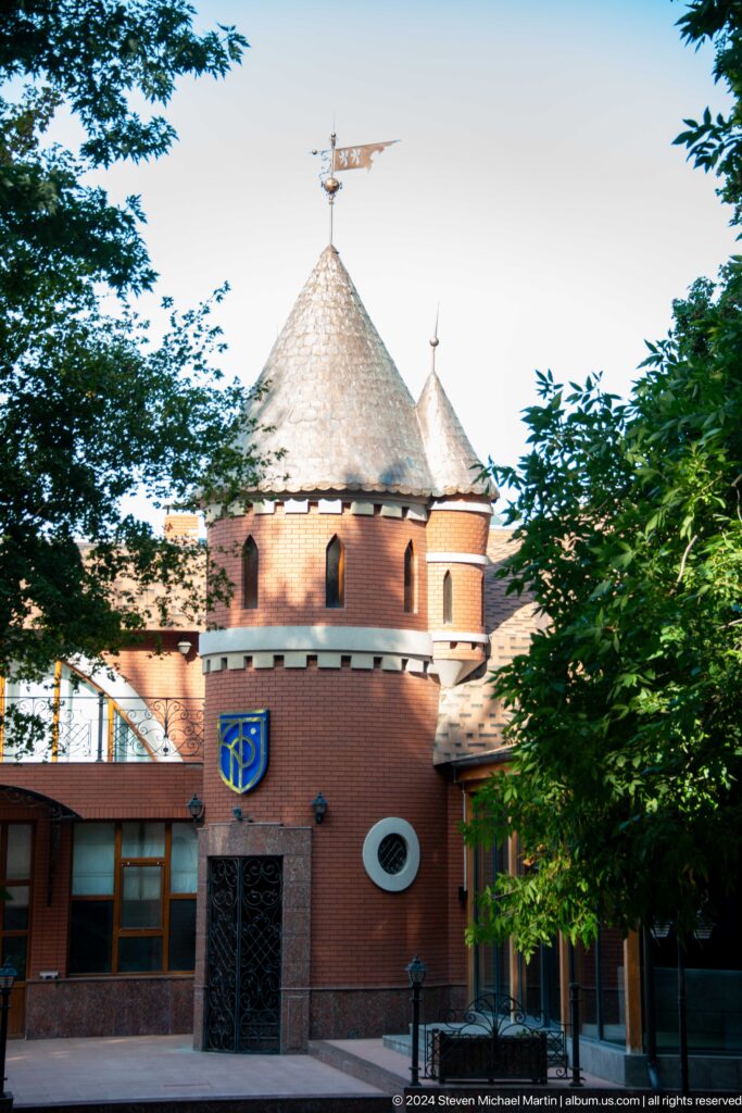 Round red-brick tower with narrow windows, steep conical roof, blue shield, and surrounding trees.