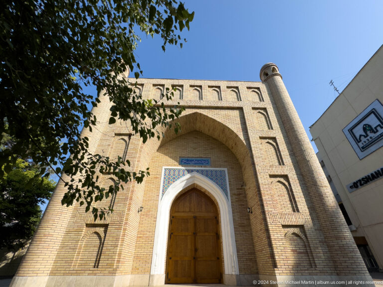 Low-angle brick mausoleum facade framed by tree branches with a modern building beside it.