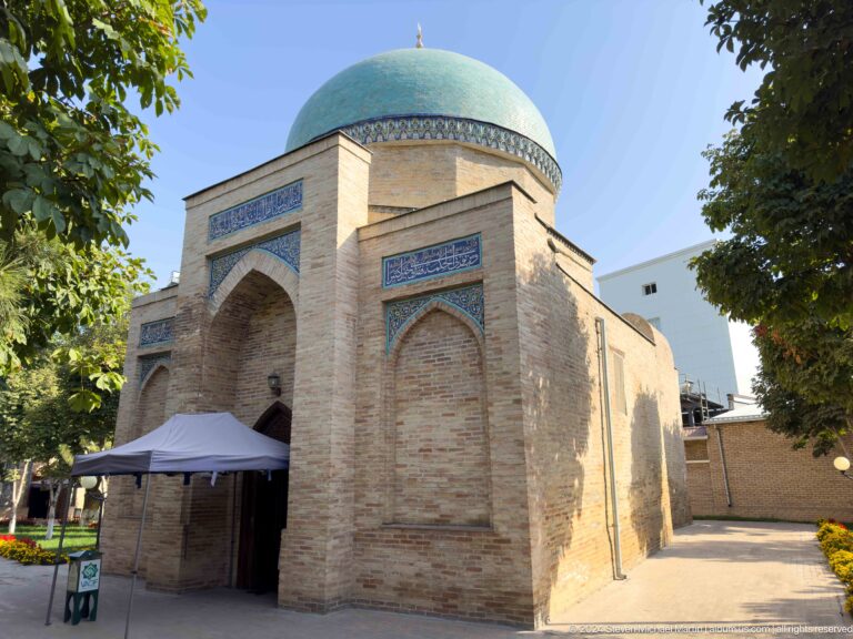 Three-quarter view of a turquoise-domed brick mausoleum with blue inscriptions, pointed niches, and entrance canopy.