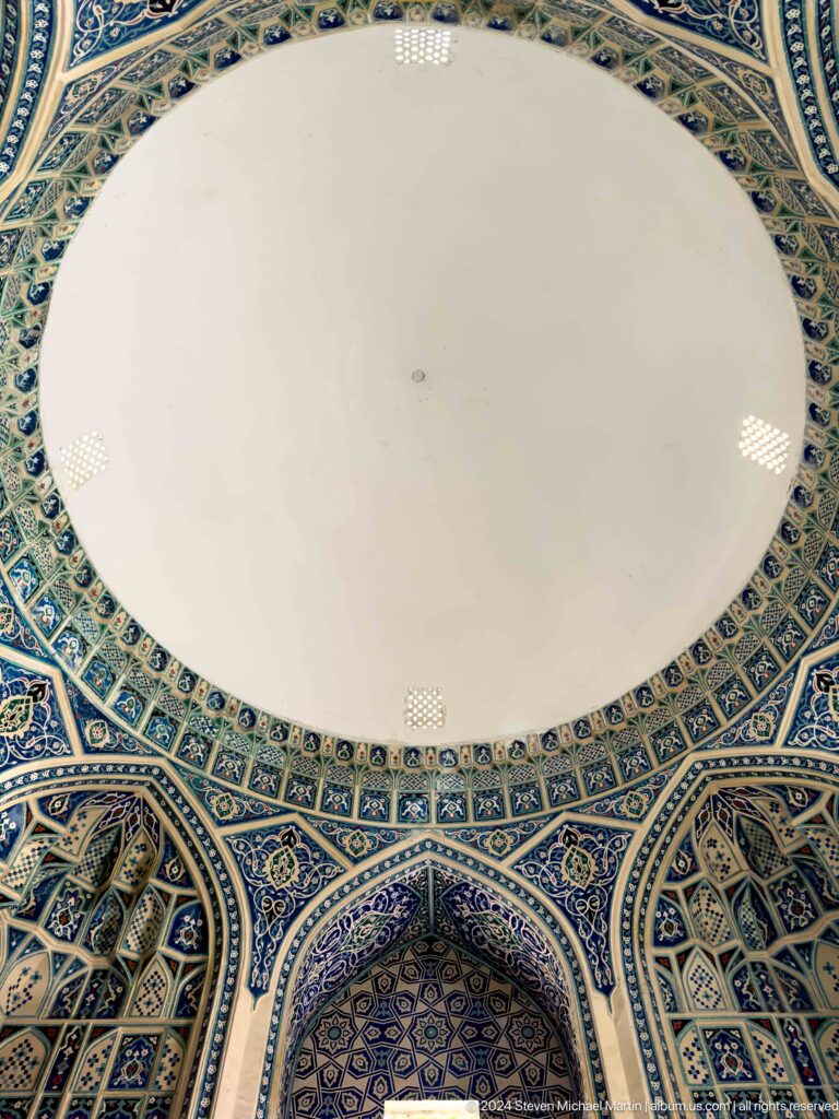 Upward view of a plain white dome bordered by blue tilework and small lattice windows.