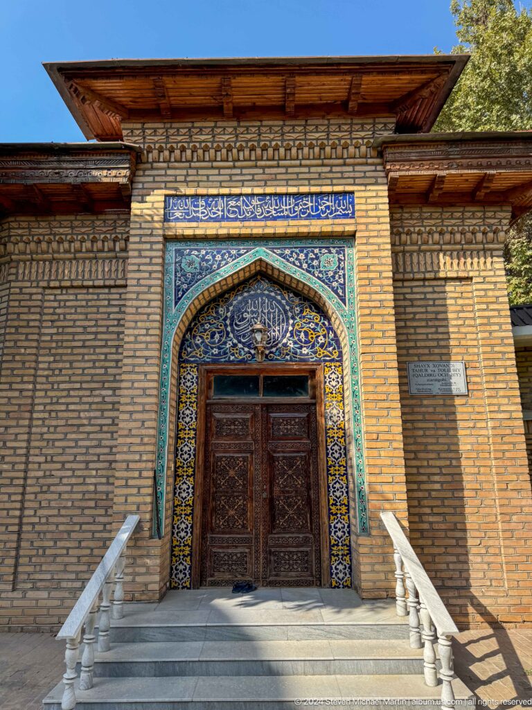 Carved wooden mausoleum door framed by blue floral tile and Arabic calligraphy, with a plaque beside it.