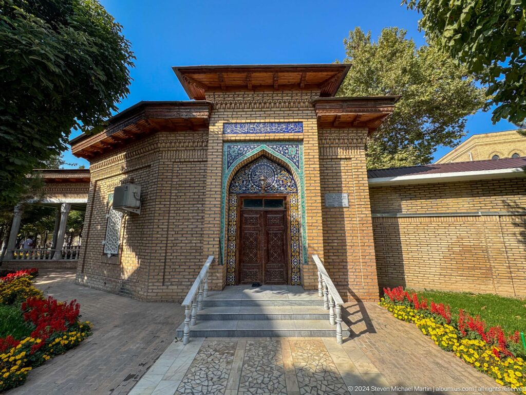 Small brick mausoleum with deep timber eaves, blue-tiled doorway, white steps, and red flower beds.