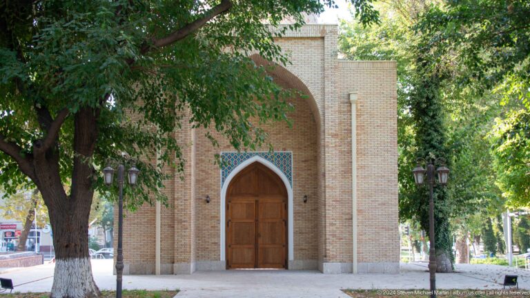 Small brick mausoleum entrance with pointed wooden door beneath a broad recessed arch and trees.