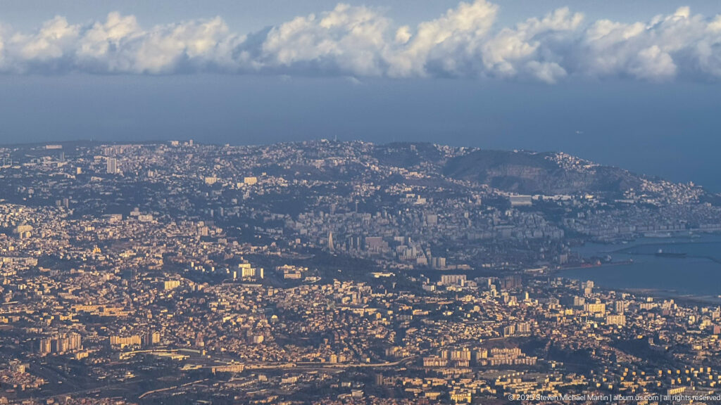 High-altitude aerial view of dense Algiers neighborhoods, hills, curving coastline, and the Mediterranean under broken clouds.