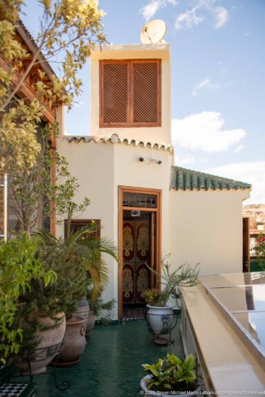 Whitewashed terrace with potted plants, decorative wooden door, timber shutters, green floor covering, and satellite dish.