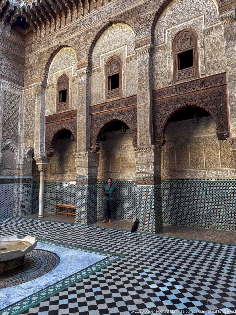 Historic madrasa courtyard with zellij, carved stucco, dark wooden galleries, patterned floor, and central basin.