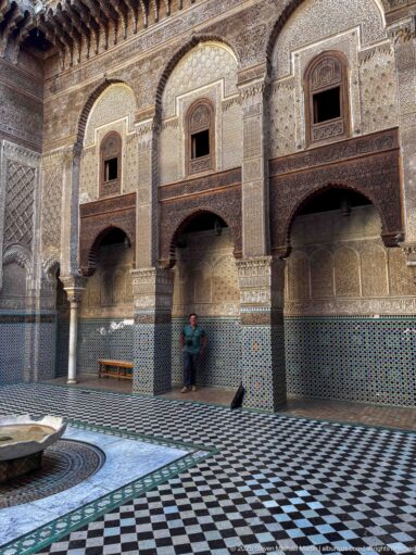 Historic madrasa courtyard with zellij, carved stucco, dark wooden galleries, patterned floor, and central basin.