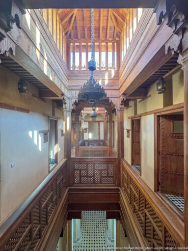Two-story wooden interior gallery with carved balustrades, doors, timber roof, and hanging lanterns around a central opening.