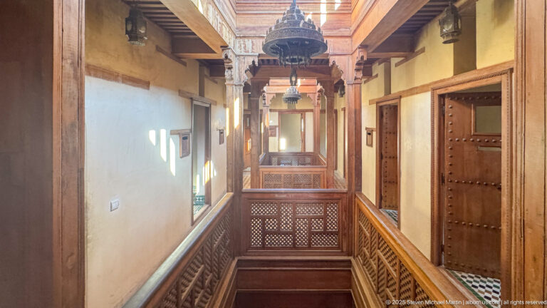 Upper-level wooden corridor with carved railings, heavy doors, hanging lanterns, and an open courtyard void.