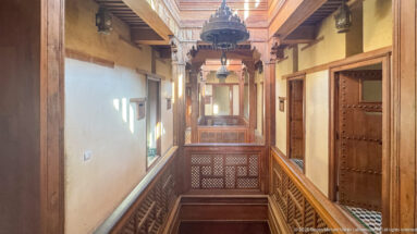 Upper-level wooden corridor with carved railings, heavy doors, hanging lanterns, and an open courtyard void.