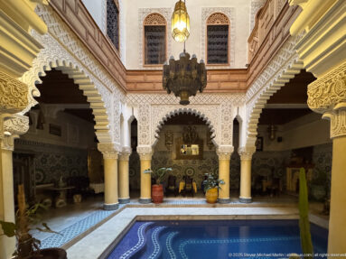 Arcaded Moroccan courtyard with blue-edged water feature, scalloped arches, columns, carved plaster, and hanging lamps.