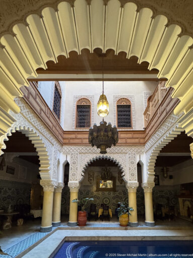 Wide courtyard view with scalloped arches, pale columns, carved plaster, wooden galleries, lanterns, and a tiled pool edge.