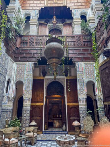 Ornate riad courtyard façade with carved wooden balconies, zellij walls, arches, greenery, and a large hanging lantern.