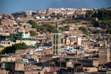 Green-tiled minaret rising among dense historic rooftops and pale masonry buildings in Fes.
