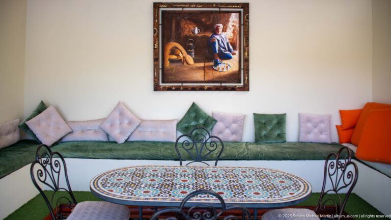 Moroccan sitting room with green banquette, pastel cushions, round mosaic table, wrought-iron chairs, and framed painting.