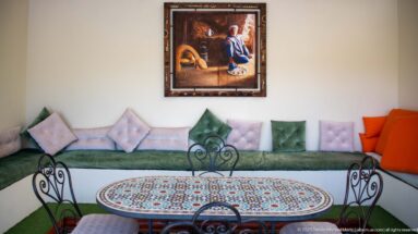 Moroccan sitting room with green banquette, pastel cushions, round mosaic table, wrought-iron chairs, and framed painting.