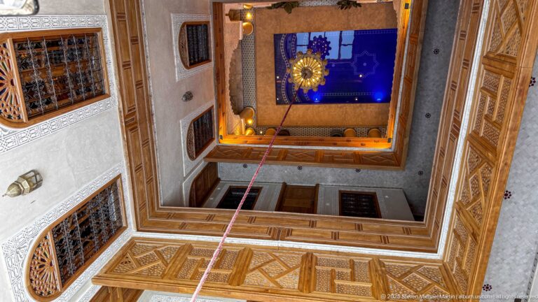 Upward view through a square riad courtyard with carved wooden galleries, screens, lantern, and blue sky.