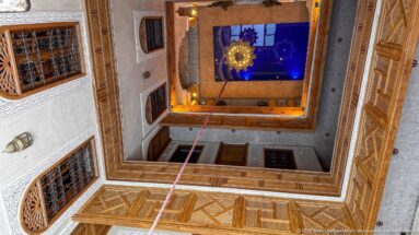 Upward view through a square riad courtyard with carved wooden galleries, screens, lantern, and blue sky.
