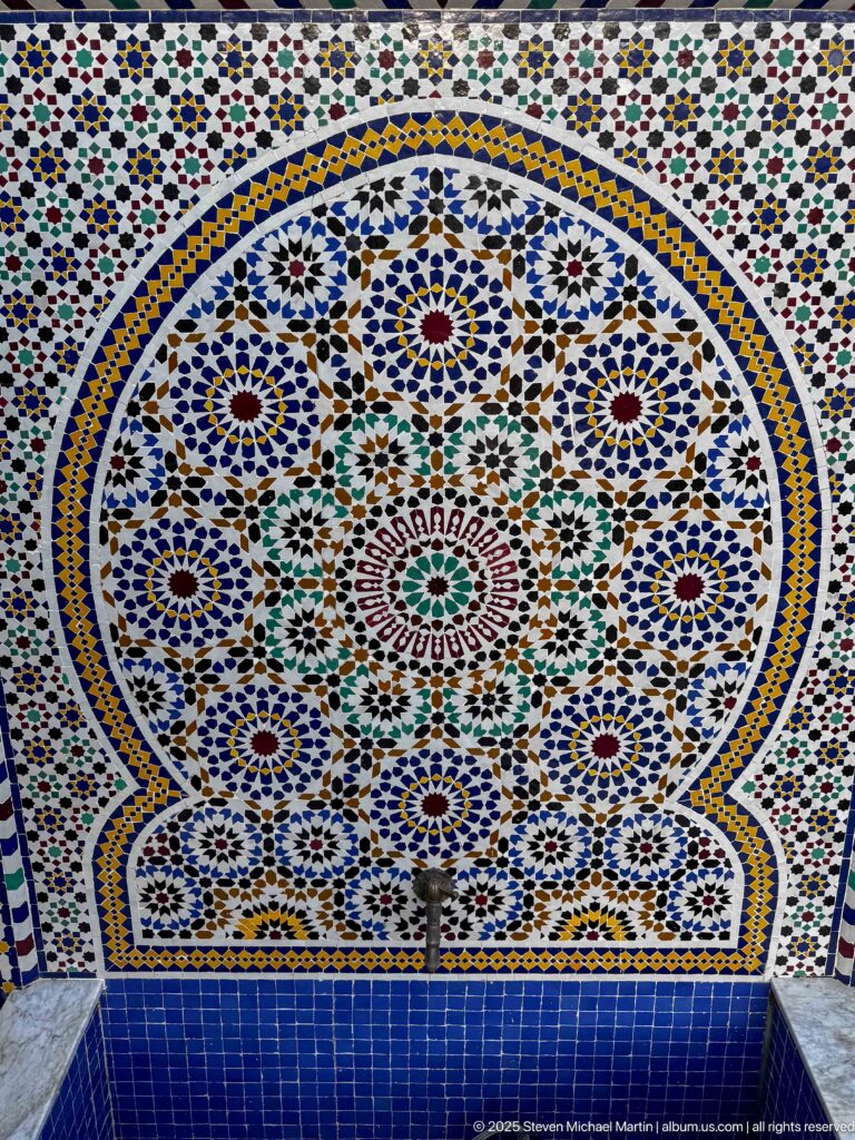 Wall fountain centered in a scalloped field of multicolored zellij with dense geometric patterns.