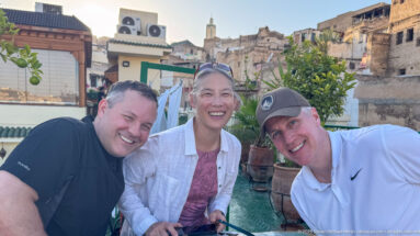 Three travelers smiling together on a rooftop terrace with old masonry buildings and a distant minaret behind them.
