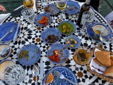 Shared Moroccan meal of small plates, bread, drinks, and condiments on a black-and-white mosaic table.