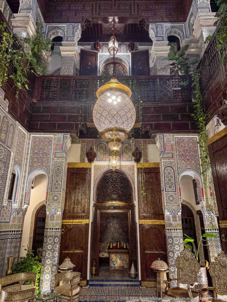 Two-story riad courtyard with a large hanging lantern, carved wooden balconies, arches, and zellij walls.