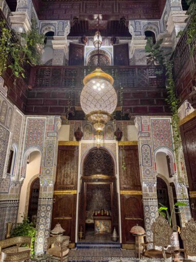 Two-story riad courtyard with a large hanging lantern, carved wooden balconies, arches, and zellij walls.