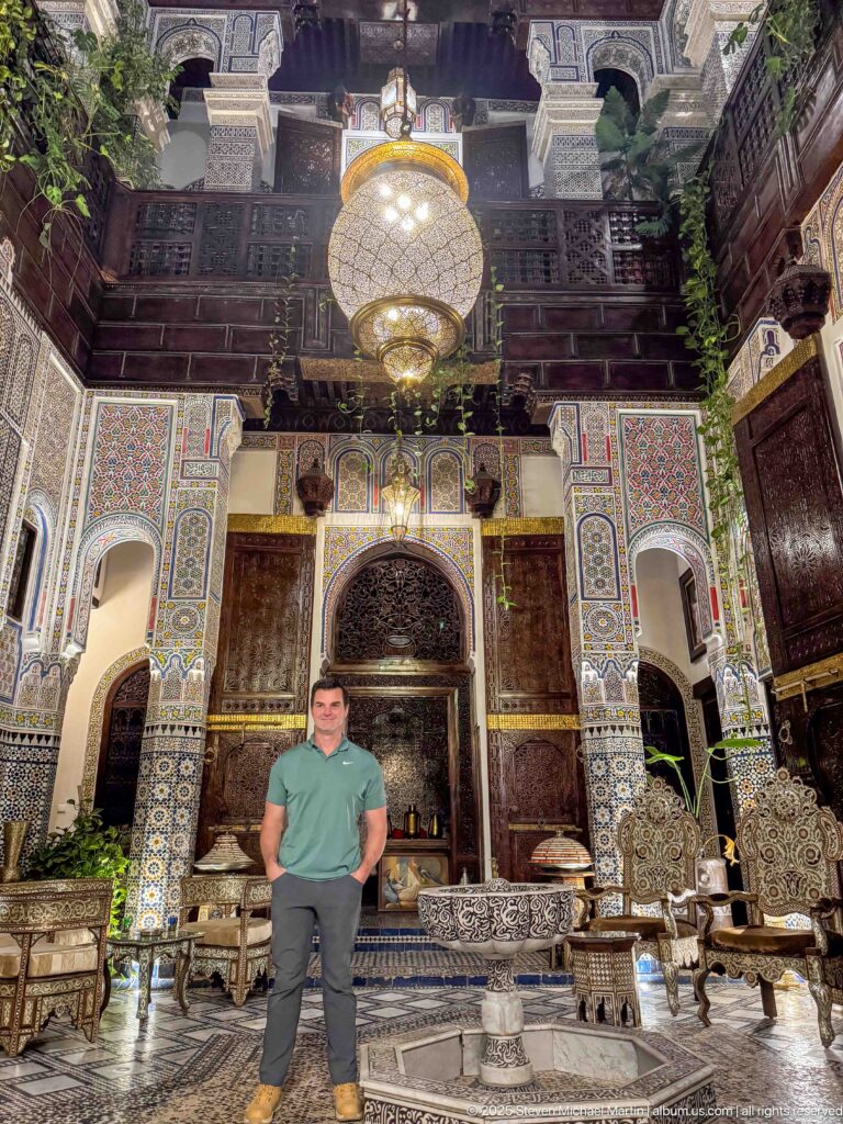 Traveler standing beneath a large hanging lantern in an ornate two-story riad courtyard.
