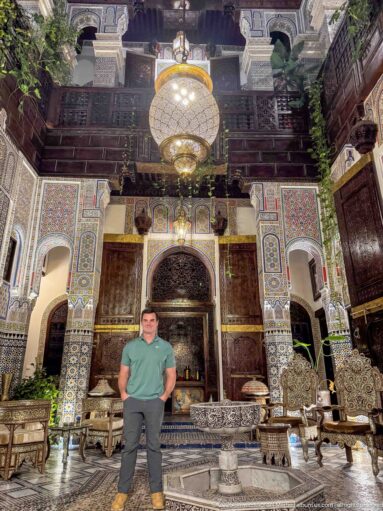 Traveler standing beneath a large hanging lantern in an ornate two-story riad courtyard.