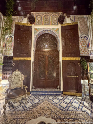 Ornate Moroccan salon with carved wooden doorway, zellij walls, patterned carpet, and traditional seating.