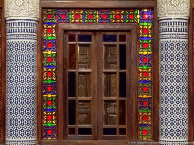 Wooden double door bordered by multicolored stained glass and blue-and-white zellij panels.