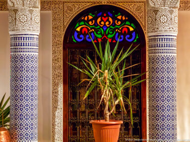 Arched stained-glass transom over a dark doorway framed by carved plaster and blue-and-white zellij.