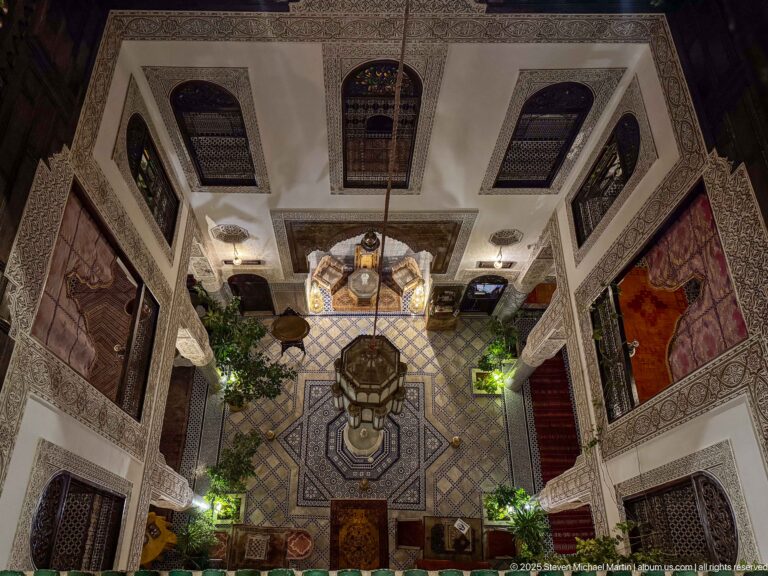 High-angle view into a multi-level riad courtyard with central fountain, patterned floor, and surrounding galleries.