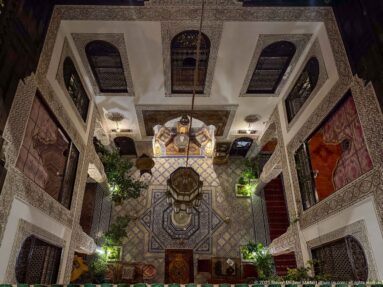 High-angle view into a multi-level riad courtyard with central fountain, patterned floor, and surrounding galleries.