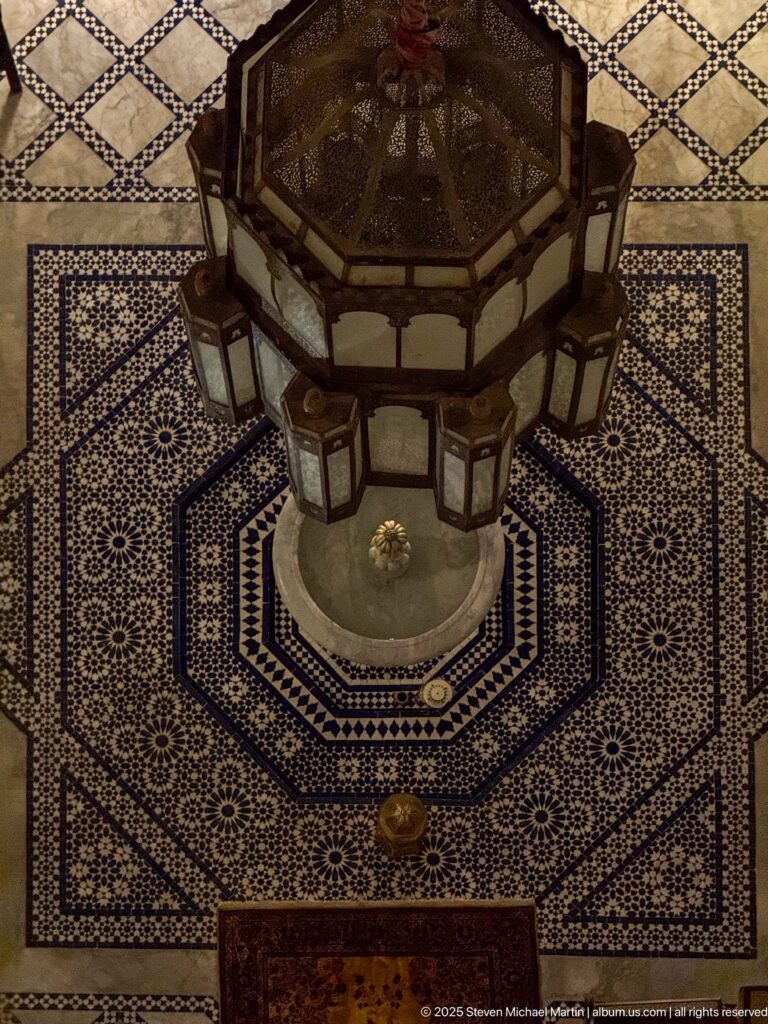 Octagonal fountain centered in a blue-and-white zellij courtyard beneath hanging lanterns.