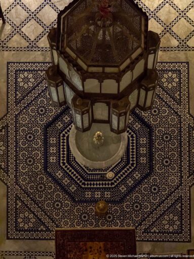 Octagonal fountain centered in a blue-and-white zellij courtyard beneath hanging lanterns.